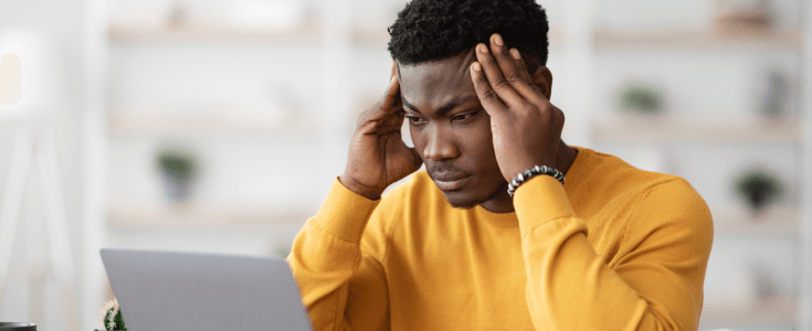 A young man sits at his laptop with his head in his hands