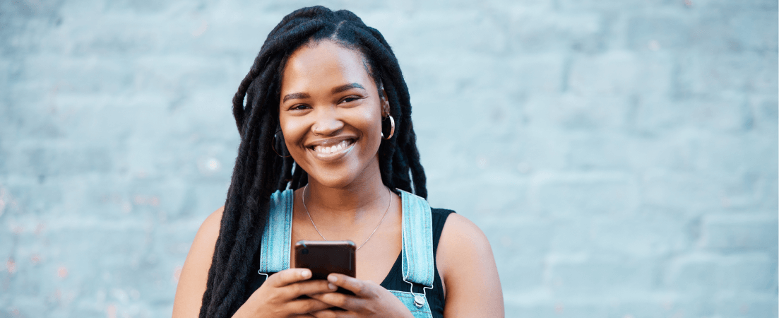 Young woman holds her phone in front of a brick wall and smiles